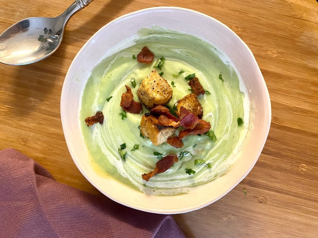 Bowl on table with creamy avocado soup topped with bacon and chile rubbed croutons.